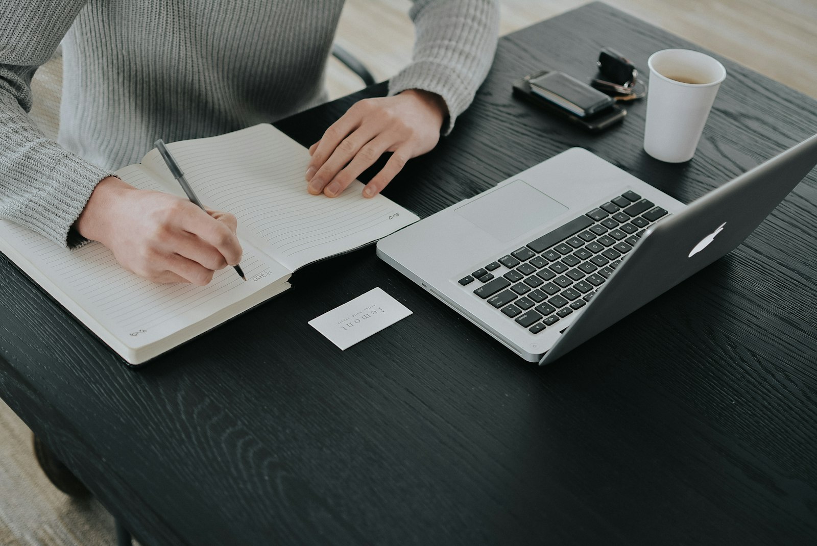 A person writing in a notebook at a dark desk, representing a new hire building a personal reference notebook during their first week