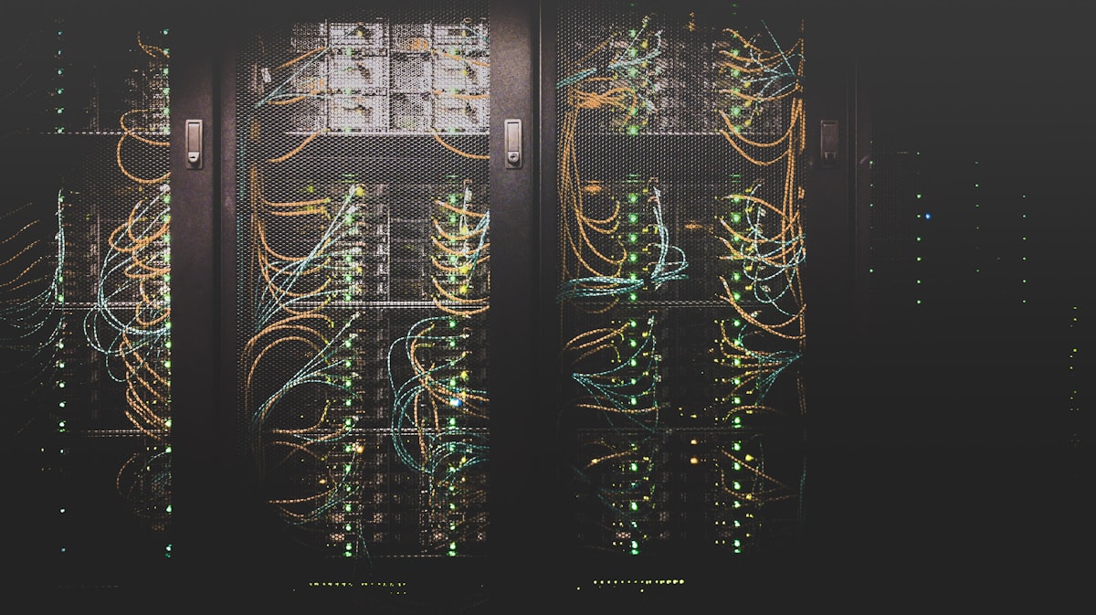 A dark server rack lit by rows of coloured status lights and cabling, representing the cloud infrastructure AI agents run on without direct human observation